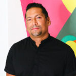 Puerto Rican man with short dark hair and a goatee, standing in front of a colorful mural. He is smiling and wearing a black button up shirt