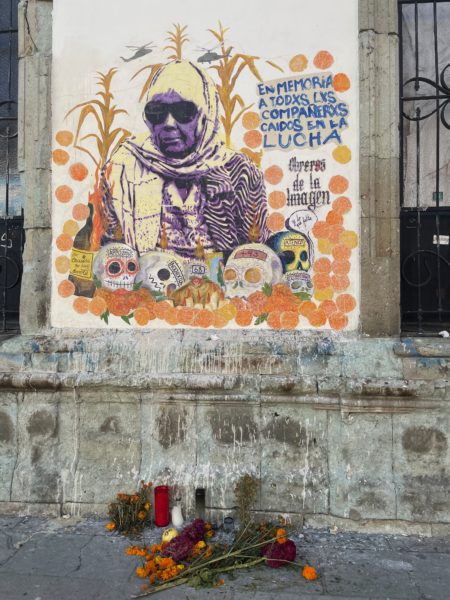 photo of a mural painted on the side of a stone building. the mural features an older woman in sunglasses, below her are various skulls and surrounding her are marigolds. below the mural are flowers and candles placed on the sidewalk.