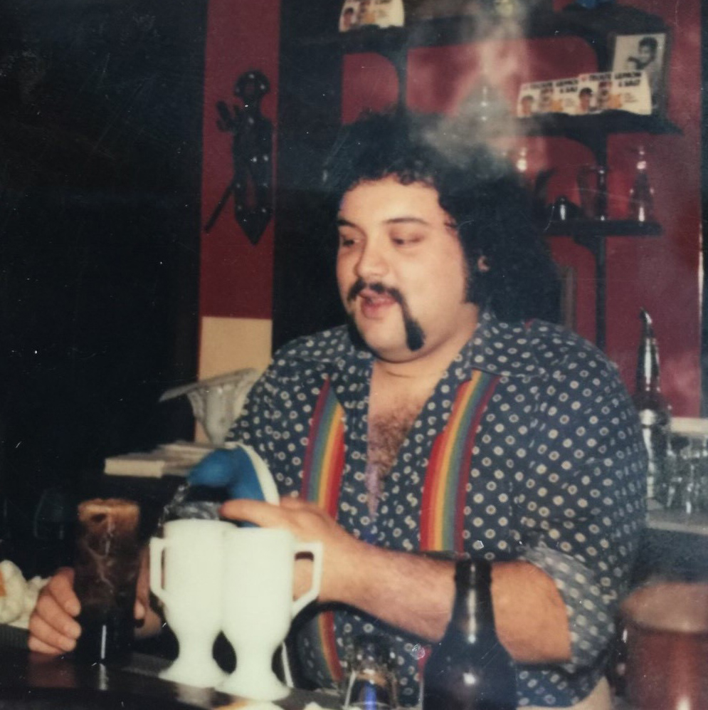 man in polka dot shirt and rainbow suspenders making drinks behind a bar