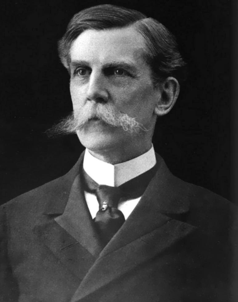 A black and white photo of a man wearing a suit and tie with hair combed over and a long, handlebar mustache. 