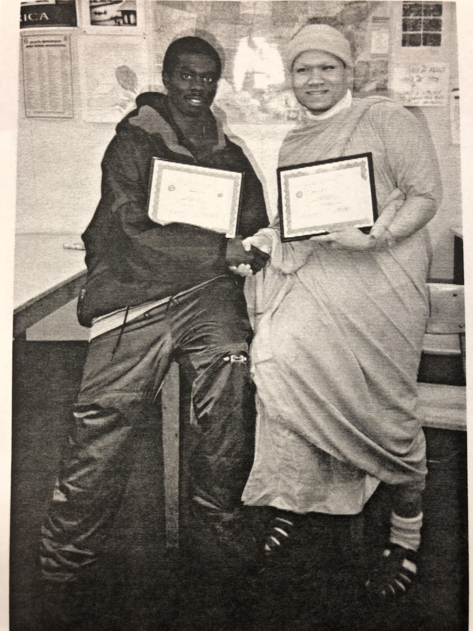 A black and White photo of two teens, one Black and one Laotian wearing traditional monk robes, both holding certificates of achievement.