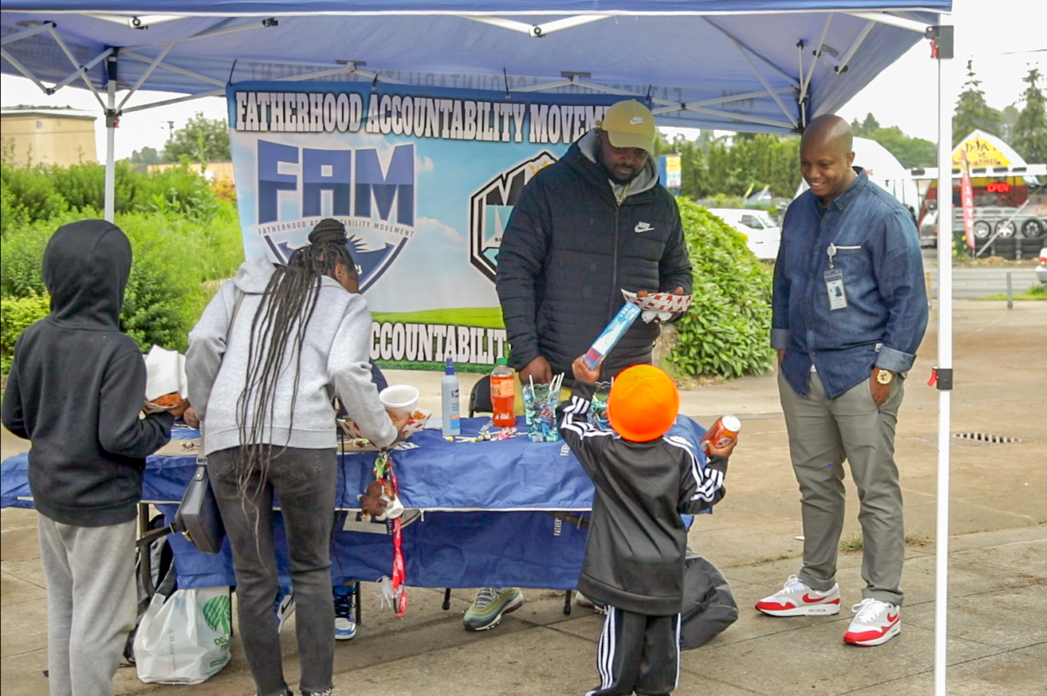 Two men stand by a table, beneath a top-up canopy at a neighborhood street fair looking down talking to a kid.