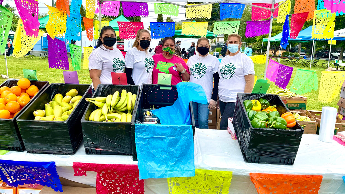 5 people in t-shirts that read "urban Fresh Food Collective" standing behind a table filled with produce at an outdoors market