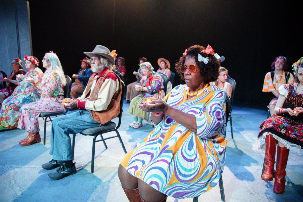 photo from a theatrical production of a diverse group of people in colorful costumes sitting in chairs on a stage
