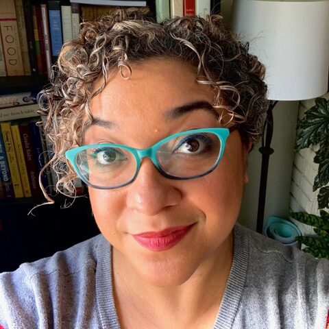 photo of a Black woman's face. There is a bookshelf full of books behind her. She as curly black and blond hair and is wearing blue glasses. She is smiling.