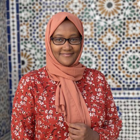 photo of a Black woman wearing a vivid floral print dress and a head scarf. She is wearing glasses and smiling at the camera.