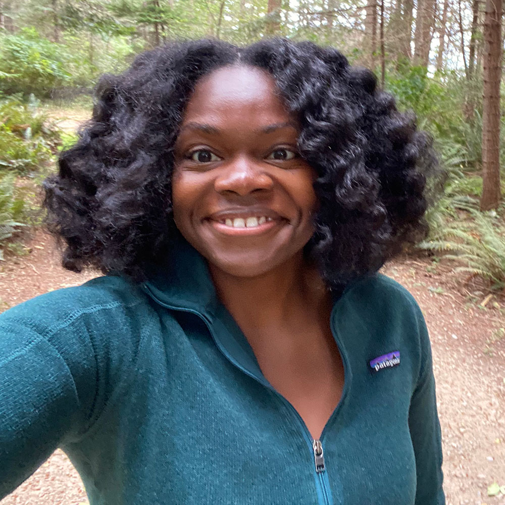 Black woman on a forest trail. She has medium length curly hair, is wearing a green fleece jacket, and smiling widely.