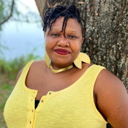 photo of Nacala Ayele, a Black woman with short braided hair. She is standing outside wearing a bright yellow dress and smiling at the camera.