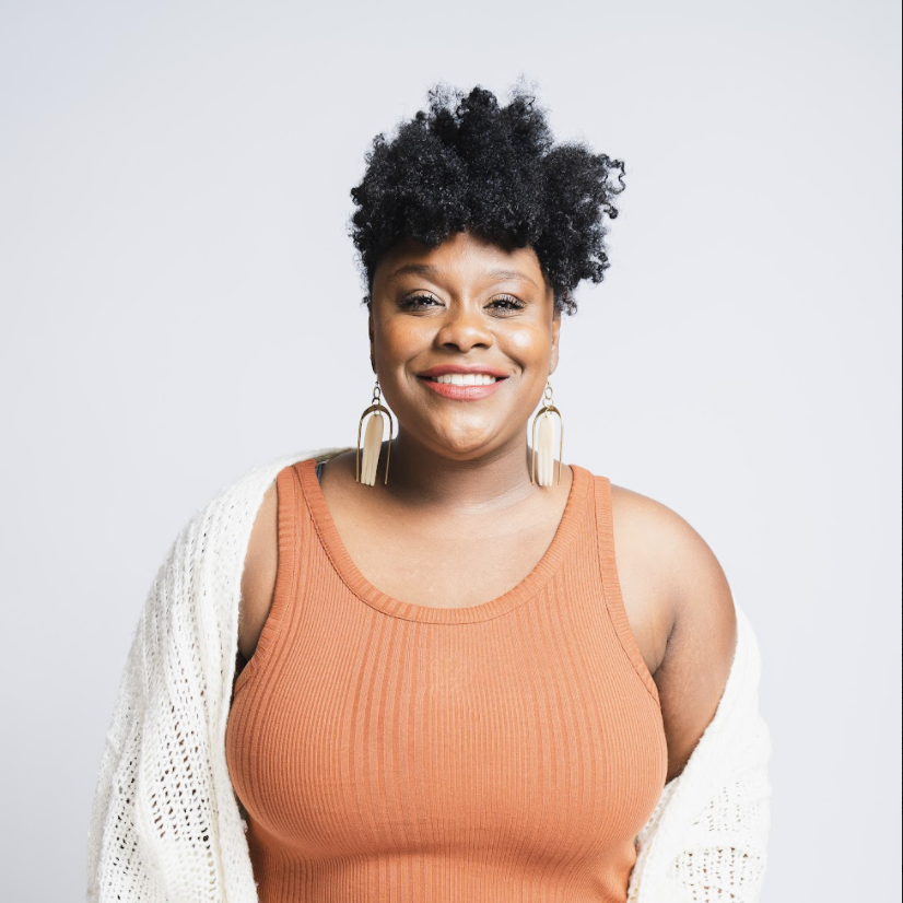 photo of a Black woman with short, curly hair standing in front of a white backdrop. she is wearing a rust colored tank top with a white sweater hanging off one shoulder. She is smiling.