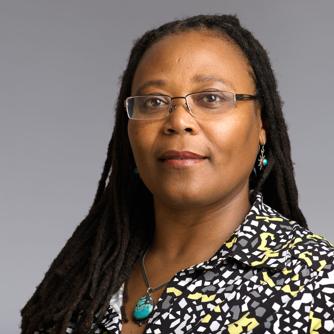 photo of Victoria Santos, an Afro-Latina woman with long dreadlocked hair. She is wearing glasses and a black blouse with yellow and grey geometric shapes.