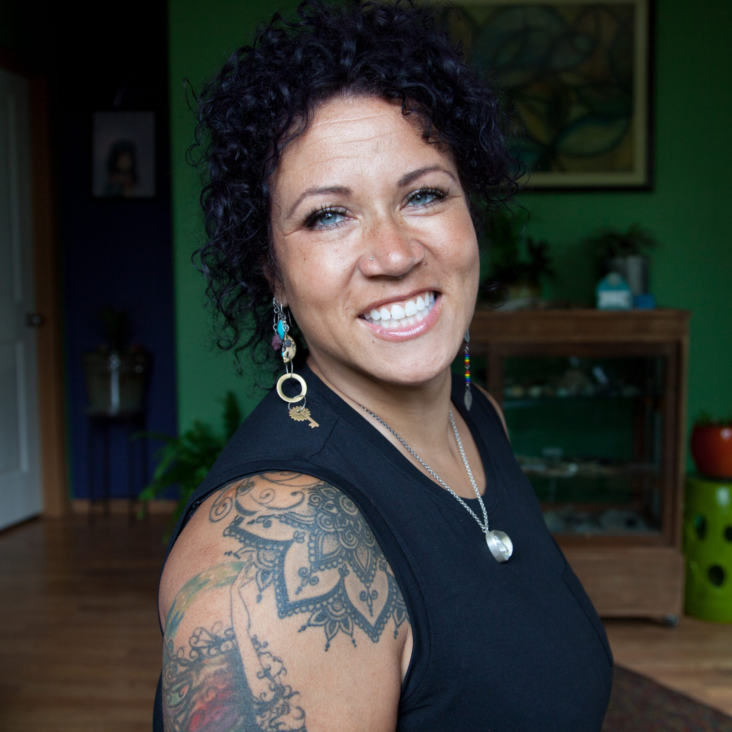 photo of a Black woman sitting in her living room. She has long curly hair and a tattooed shoulder and bicep. She is smiling at the camera.