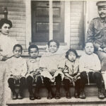 archival photo of a family on a front porch. A Filipino woman in a white dress and a Black man in military uniform stand on either side of five Black-Filipino children who are seated on a step.