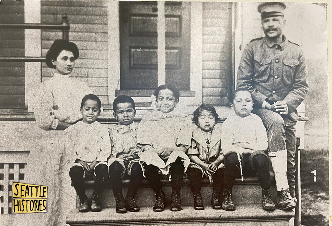 archival photo of a family on a front porch. A Filipino woman in a white dress and a Black man in military uniform stand on either side of five Black-Filipino children who are seated on a step.