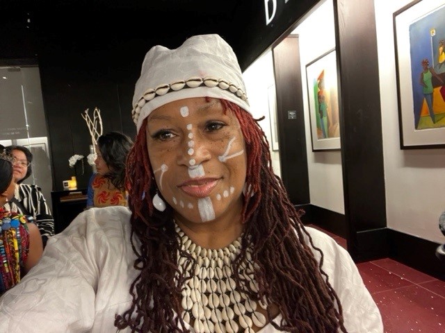 A Black woman with long braids dyed slightly red, wearing a white cap with a band of shells and markings on her face with white paint. 