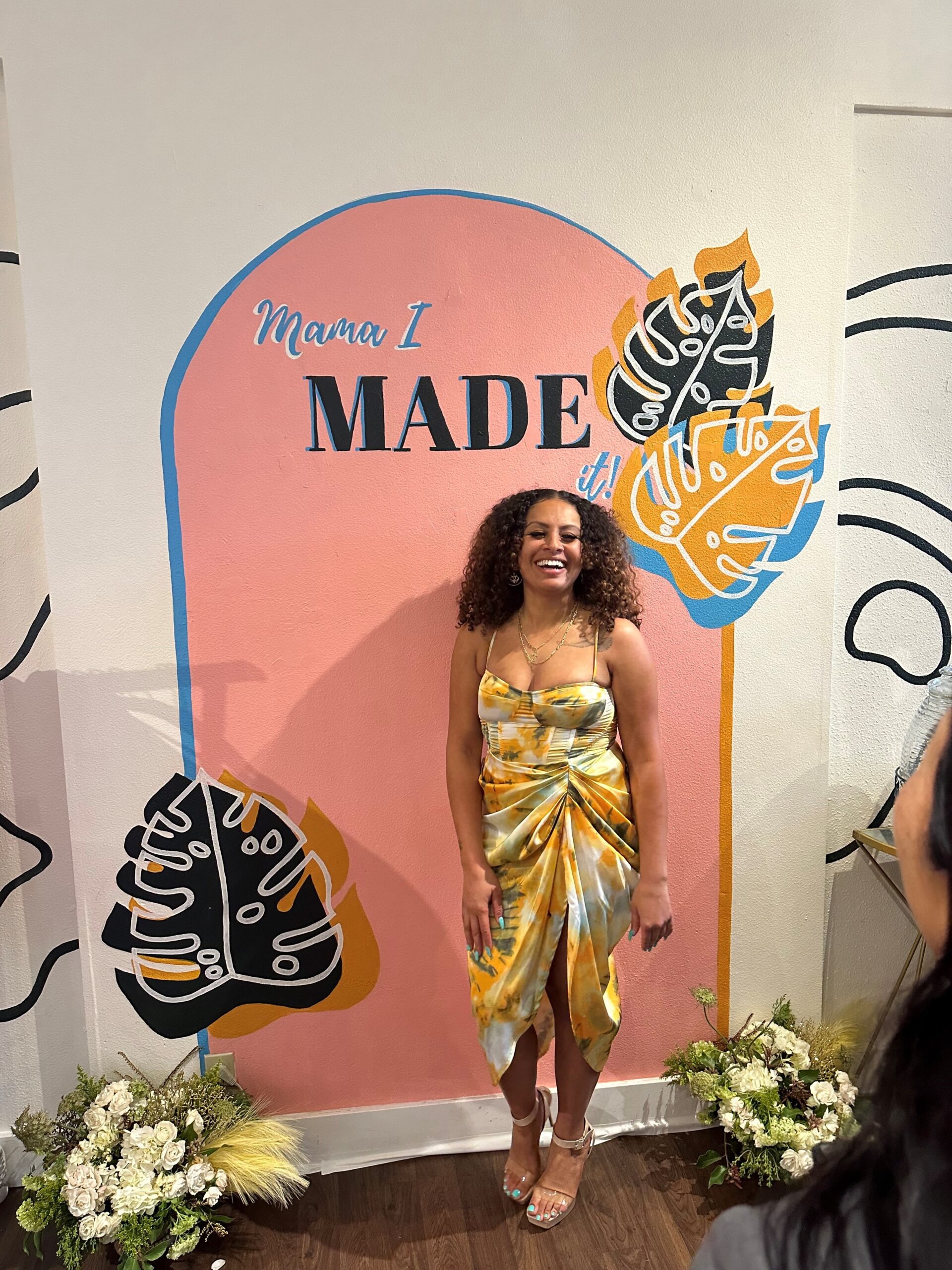 A Black woman wearing a yellow dress and heals stands in front of a sign with painted fern leaves and text that reads "Mama I MADE it!" 