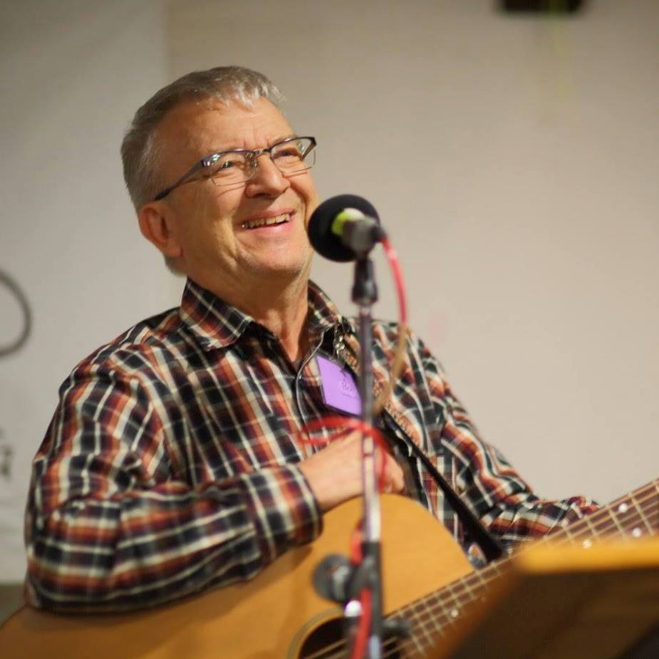 An older white man with short hair and glasses, smiling, holding a guitar and standing behind a microphone. 