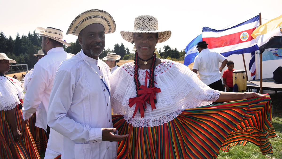 An Afr0-Latino man and woman standing outside, smiling at the camera. They are obviously performers - the man is wearing a white button-up shirt and hat; the woman is wearing a white blouse and hat, with a multicolored striped skirt.