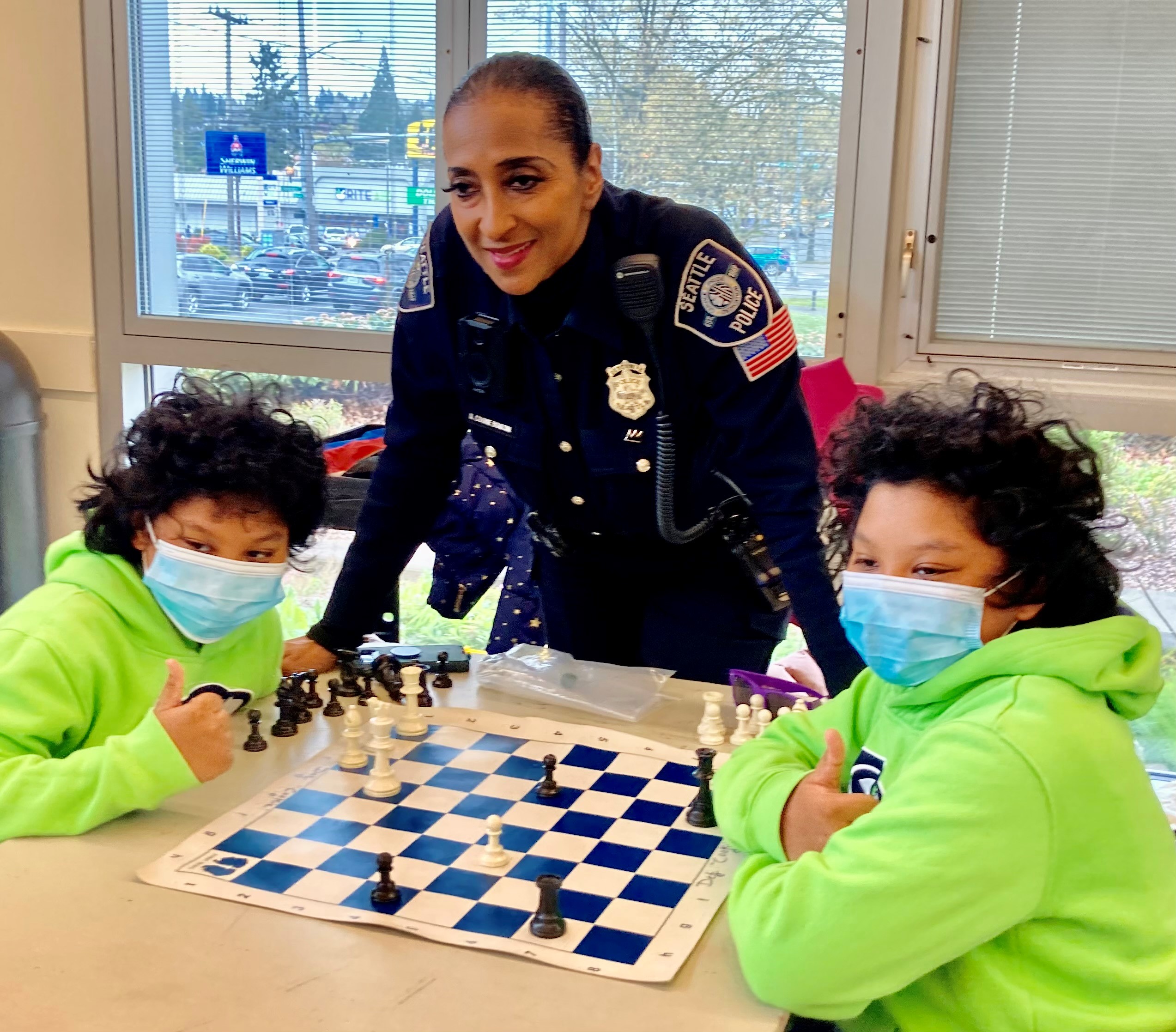 A Black woman wearing her police uniform stands at a table between two young children playing chess.