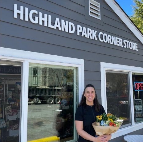 A woman standing and holding flowers outside a building with gray siding and lettering that reads "Highland Park Corner Store."
