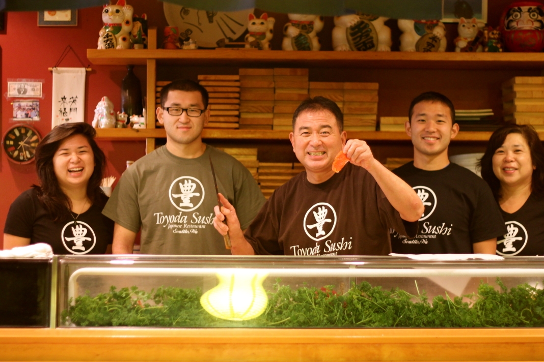 Five people stand behind a food counter smiling and all wearing shirts that say "Toyoda Sushi."