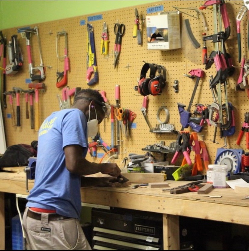 A person standing with their back to camera and wearing safety gears works at a table next to a variety of tools hanging on a peg wall. 