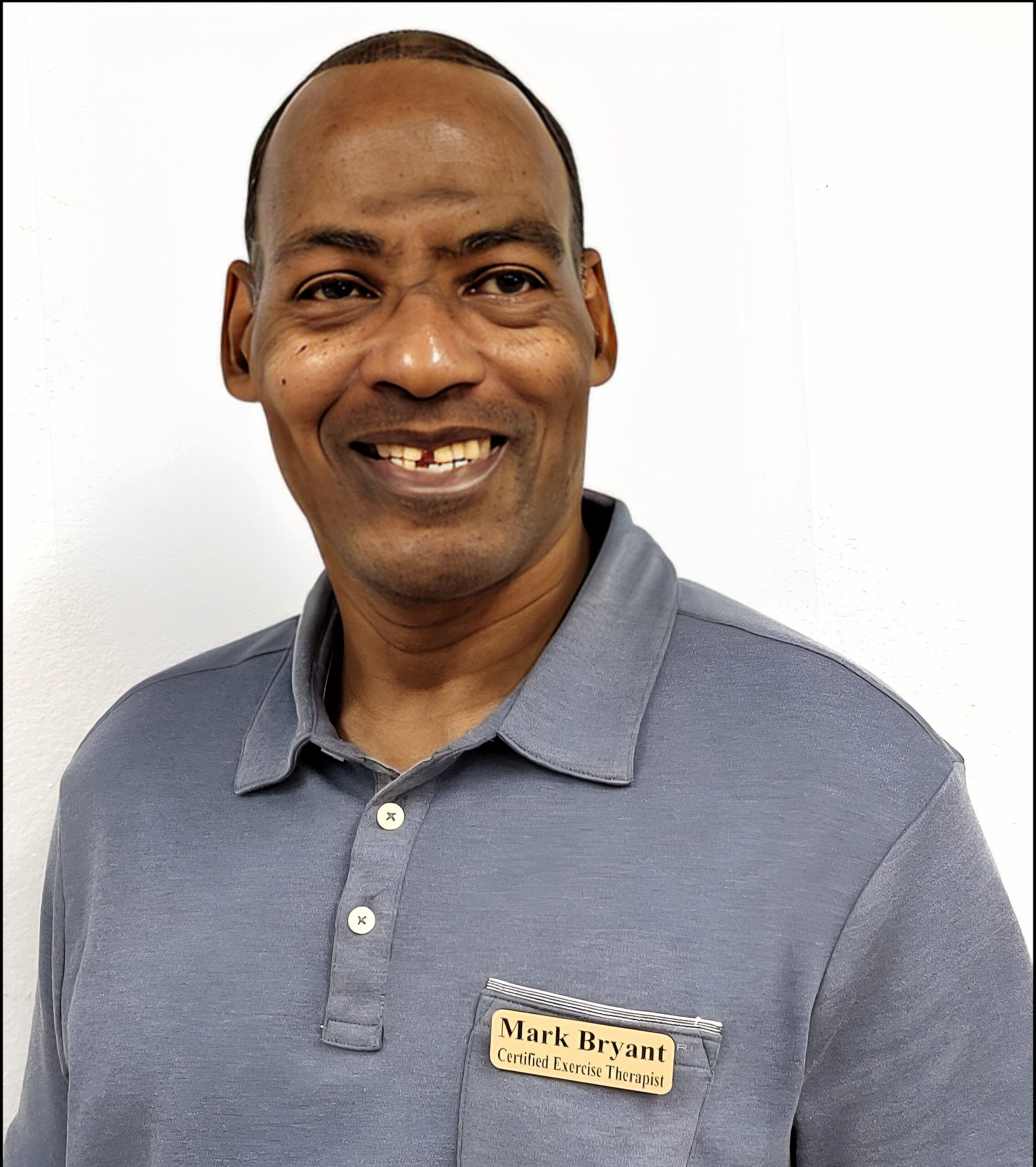 Headshot of a Black man with tight buzzed hair smiling and wearing a blue/gray polo shirt with a nametag that reads "Mark Bryant Certified Exercise Therapist"