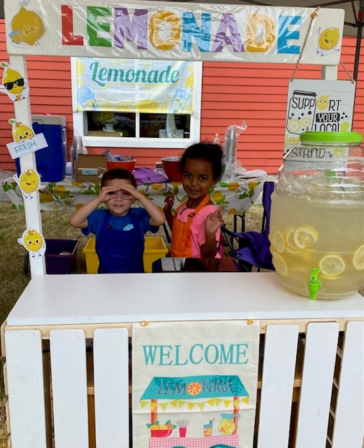 Two pre-k aged children stand at a Lemonade stand with a large container of lemonade and colorful decorations.