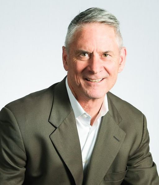 Headshot of an older white male with gray hair a white button up shirt and a brown suit coat.