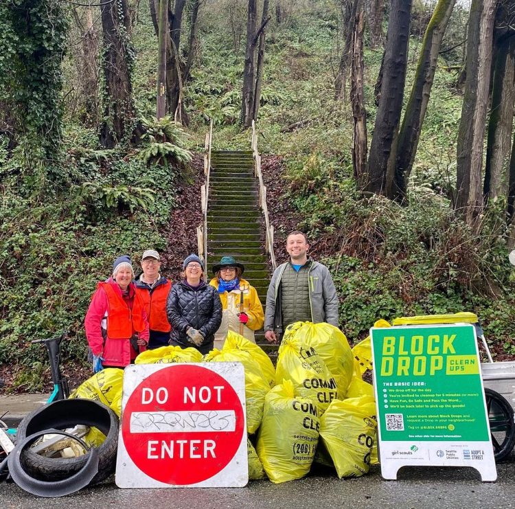 Five people standing behind a pile of full yellow garbage bags with a sign that reads "Block Drop Clean Ups."