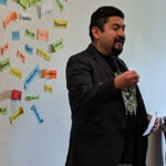 photo of Latino man standing and speaking. Behind him are many colorful pieces of paper taped to the wall with positive words like "joyous, excellence, thankful, supportive, strong", etc.