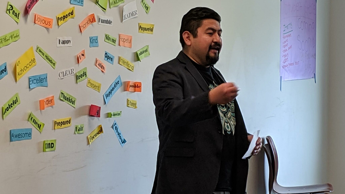 photo of Latino man standing and speaking. Behind him are many colorful pieces of paper taped to the wall with positive words like 