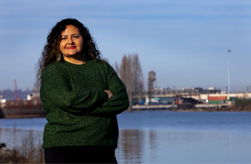 A Latinx woman with arms crossed wearing a green sweater standing at twilight with a river in the background. 
