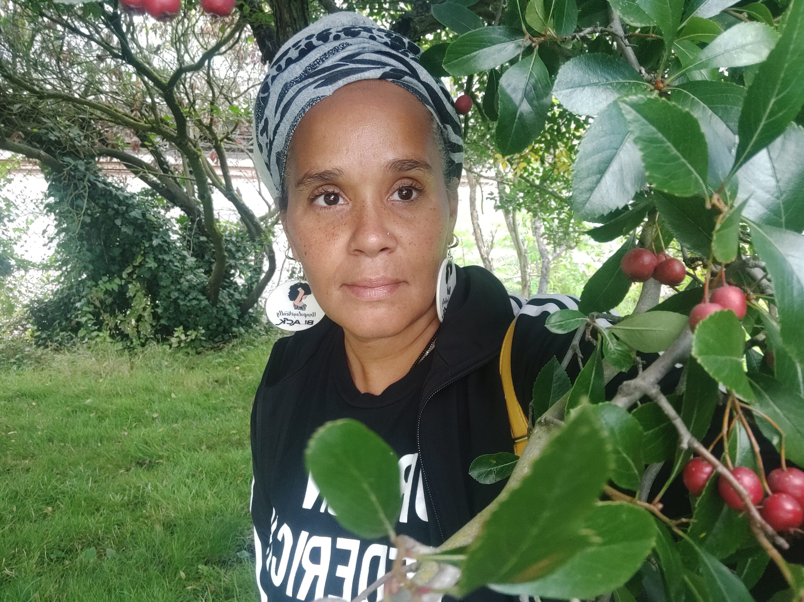 A Black woman wearing a black and white headscarf, long round earrings and a black sweatshirt standing near a tree with lush green leaves. 