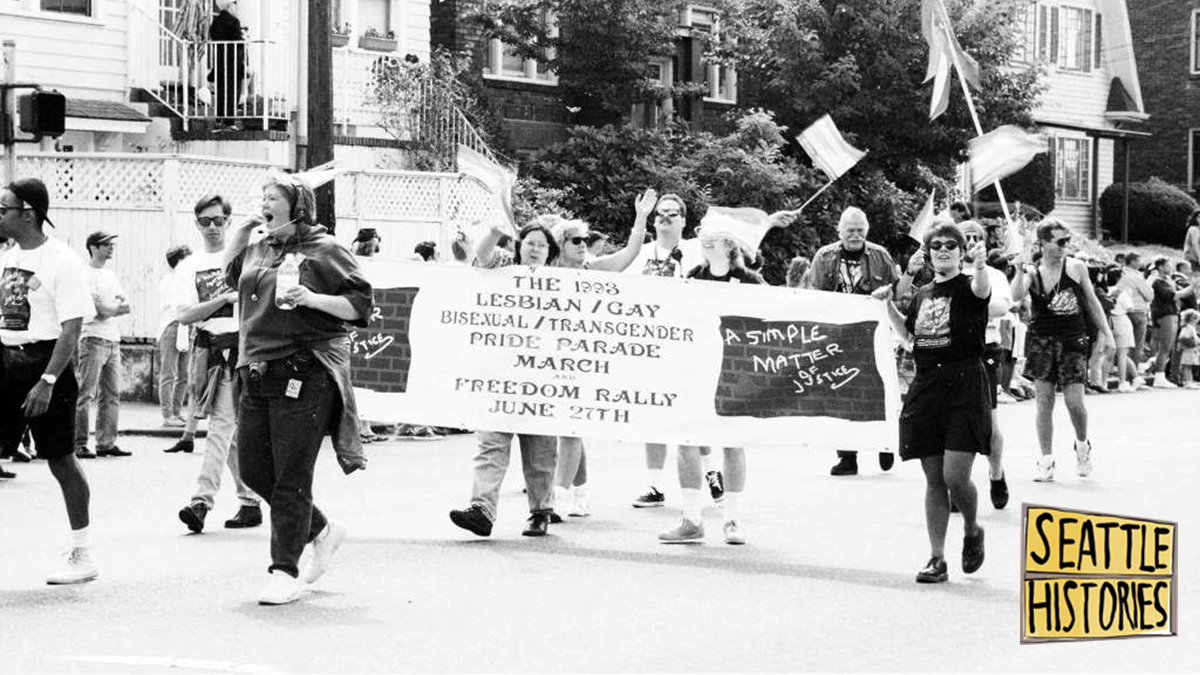 black and white photo of group of people marching in Pride Parade. several people are carrying a banner that reads 