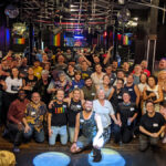 A group of 40-50 smiling people wearing nametags and posing for a photo inside a bar with a mirror on the wall, a disco ball, and a drawing of a muscular man over the words “The Cuff” on the wall behind them.