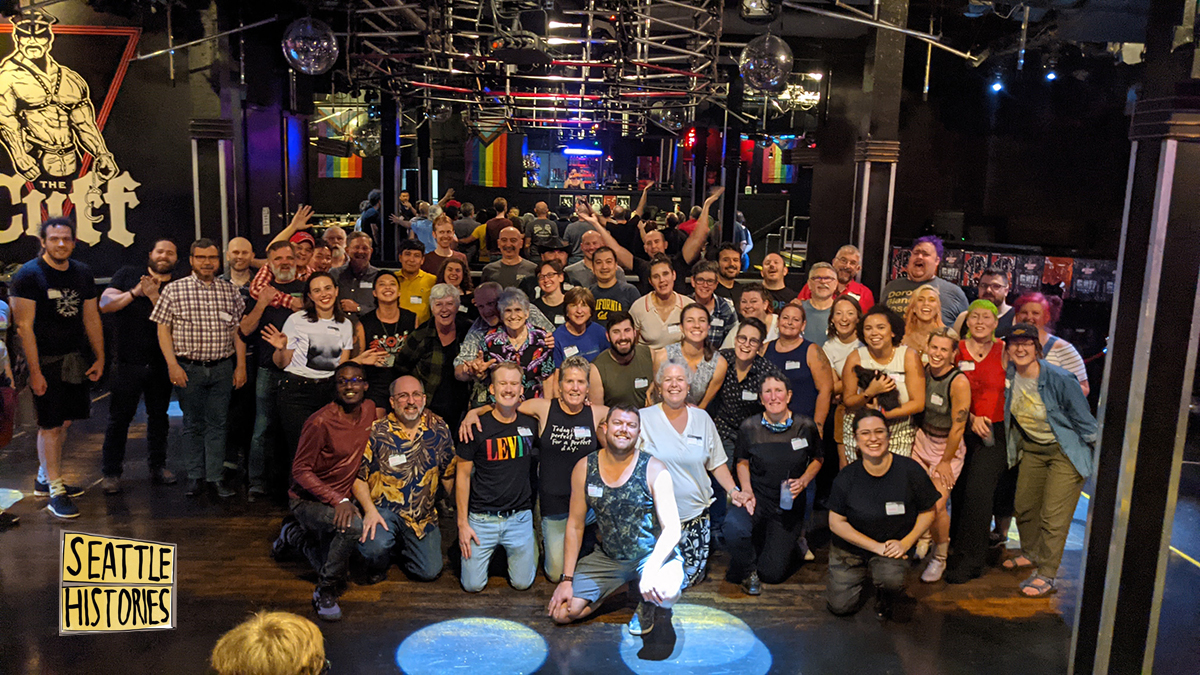A group of 40-50 smiling people wearing nametags and posing for a photo inside a bar with a mirror on the wall, a disco ball, and a drawing of a muscular man over the words “The Cuff” on the wall behind them.