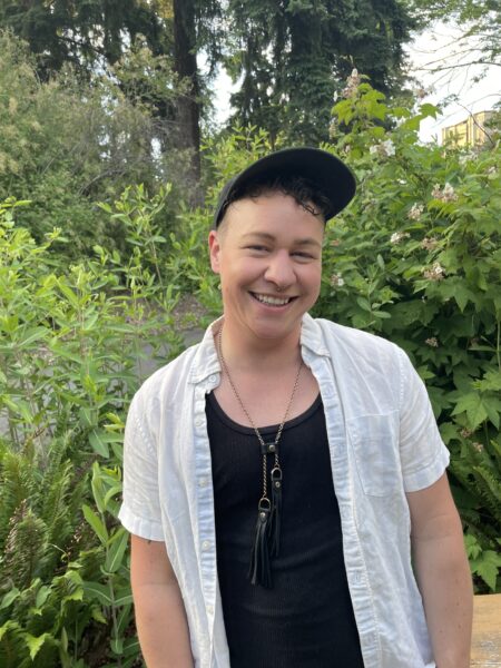 a white person smiling and wearing a black hat, short-sleeve white shirt, and a black tank top standing outside in front of greenery and trees.