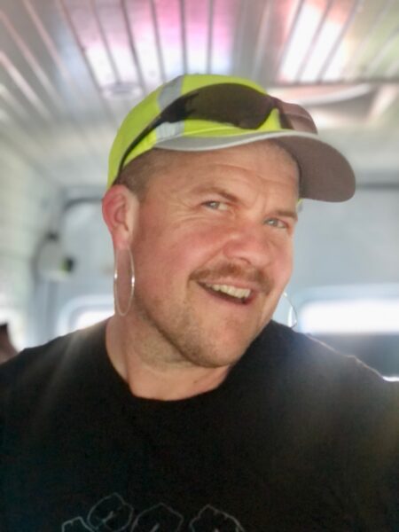 A headshot of a white person with a mustache smiling playfully and wearing a hoop earring, black t-shirt, and neon yellow ball cap with black sunglasses on the brim standing inside a small room.