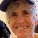 headshot of an older white woman. she has short grey hair and is smiling and wearing a cap.