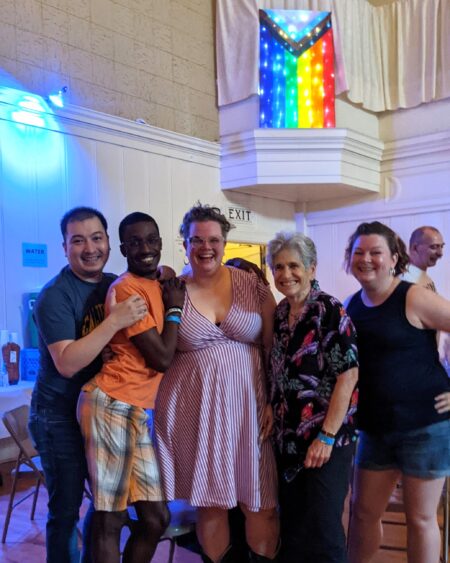 A group of five people smile for the camera in a large hall with a lit-up Pride rainbow flag behind them.