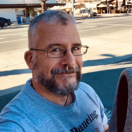 a white person with short-cropped graying hair and a cropped beard smiling outdoors in front of a paved street and wearing glasses, a septum nose piercing, and a blue t-shirt.