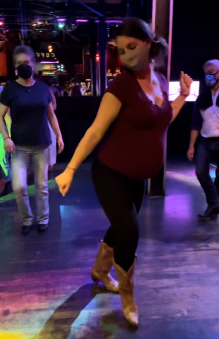 A visibly pregnant white woman in a maroon shirt, black leggings, and cowboy boots stepping forward in the midst of a dance inside on a low-lit bar dance floor, surrounded by other dancers.
