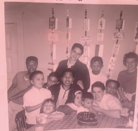 old black and white photograph of multi-racial group of young kids and two adults gathered around a table and birthday cake.