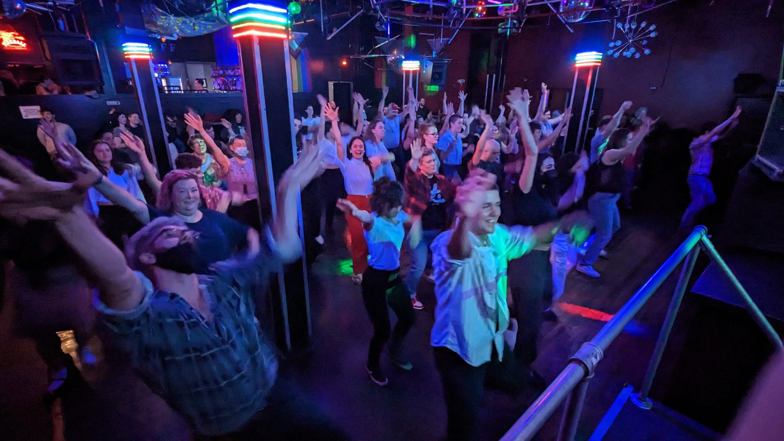 A group of approximately 35 people with their hands in the air and smiles on their faces on a dimly lit dance floor interspersed with columns wrapped in rainbow neon lights.