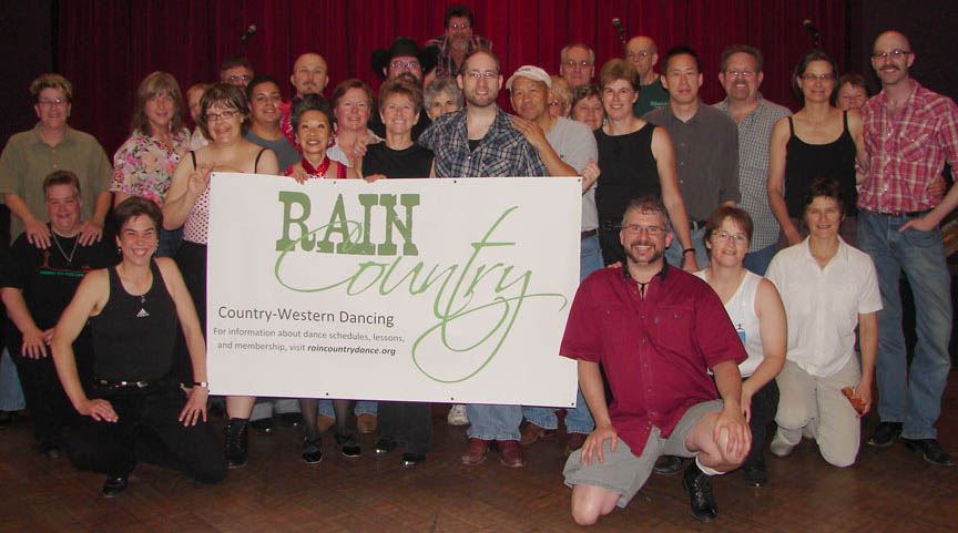 A group of twenty-nine smiling people posing for the camera and holding a white banner with green text reading “Rain Country Country-Western Dancing.”