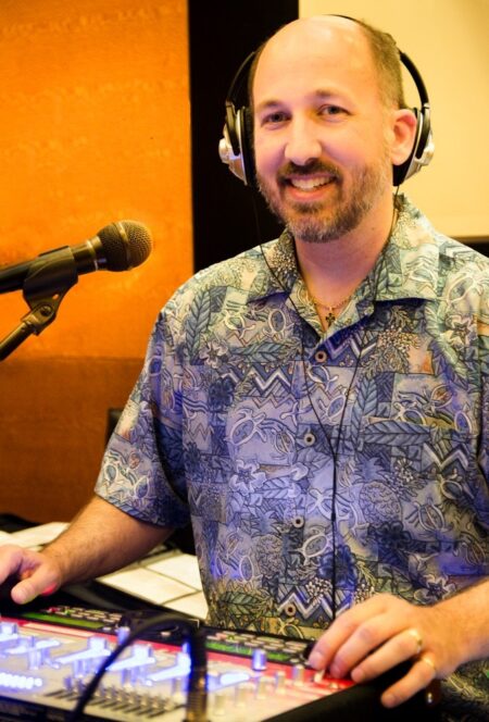 middle aged white man with beard and moustache. He is smiling, wearing headphones, and standing in front of an audio mixing board.