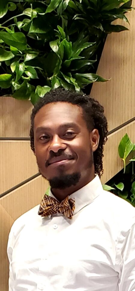 A smiling Black man with a mustache and goatee wearing a tan-patterned bow tie and white button-up shirt standing indoors in front of greenery and beige wall panels.