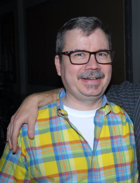middle aged white man in a plaid button-up shirt and glasses. he has a moustache and is smiling at the camera.