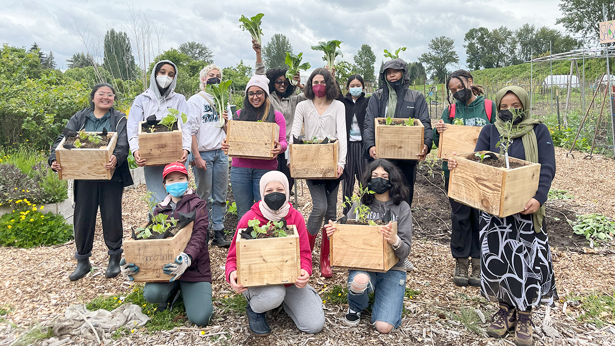 A group of 13 young women holding boxes with plants and garden greens standing in a community garden.