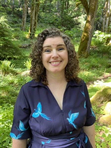 A smiling white woman with curly, shoulder-length hair wearing blue eyeliner and a dark-blue collared dress with a light-blue bird pattern standing outdoors in front of moss-covered trees, ferns, and sunlit greenery.